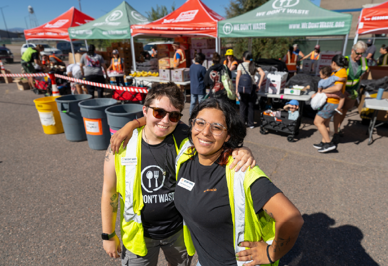 Two people with their arms around each other's shoulders in front of tents giving out food