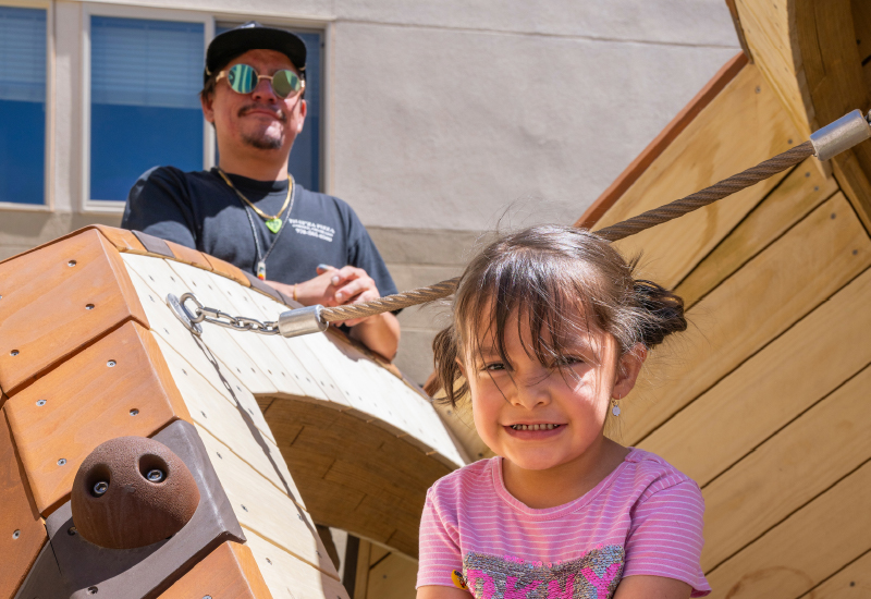 A parent smiling behind a child on a playground.