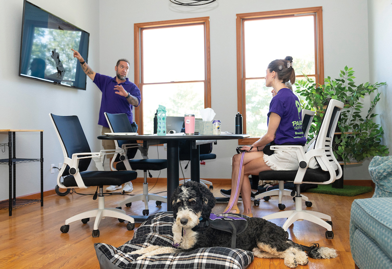 A person pointing to a TV screen, while someone listens with their service dog lying next to them.