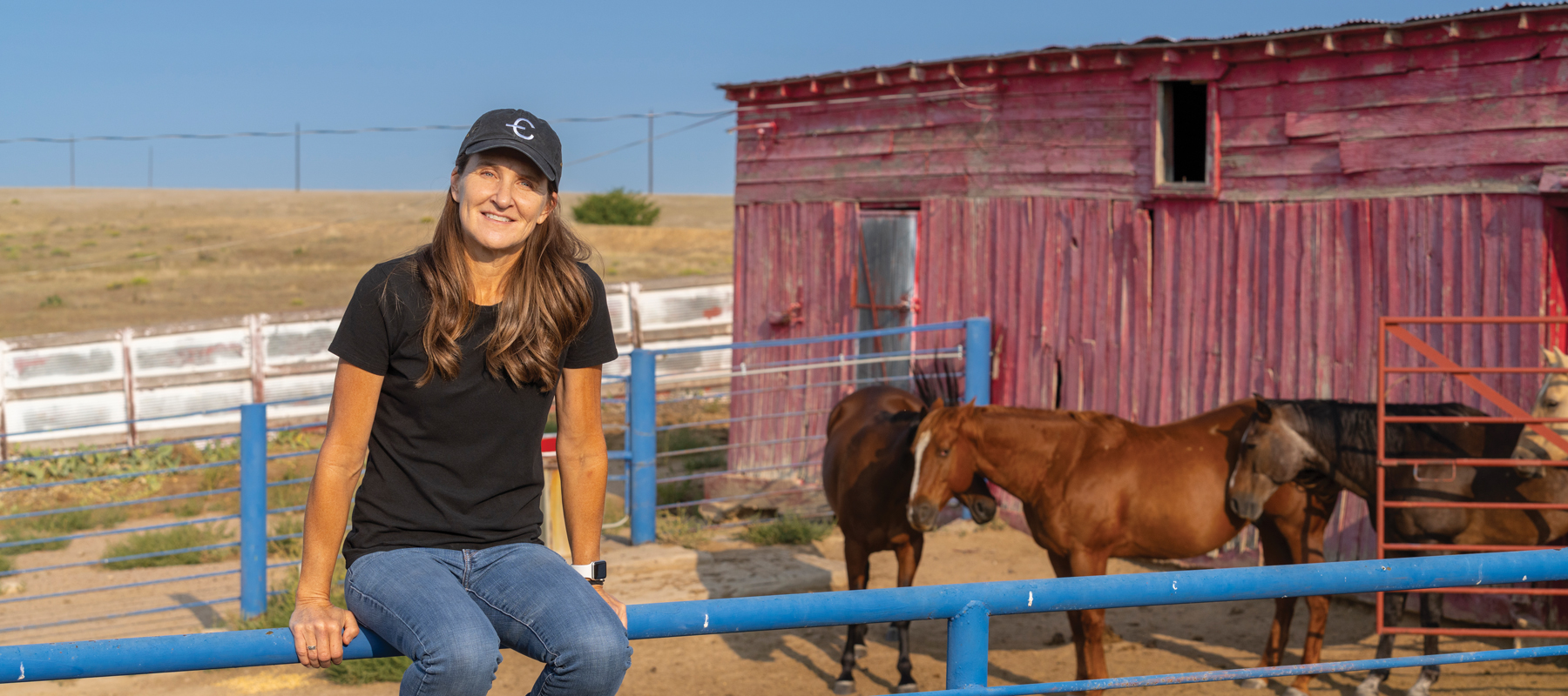 Amy Johnson sitting on a blue fence in front of a red barn and horses
