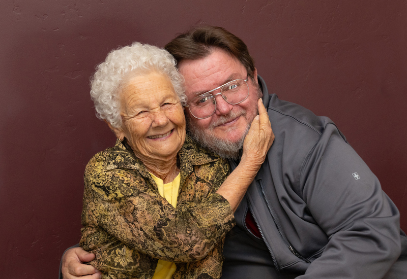 Two people in front of a maroon wall smiling for the camera, with one person holding a hand on the other's cheek