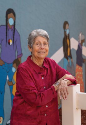 Lucille Echohawk in a red shirt leaning on a white fence with a Native American mural behind her