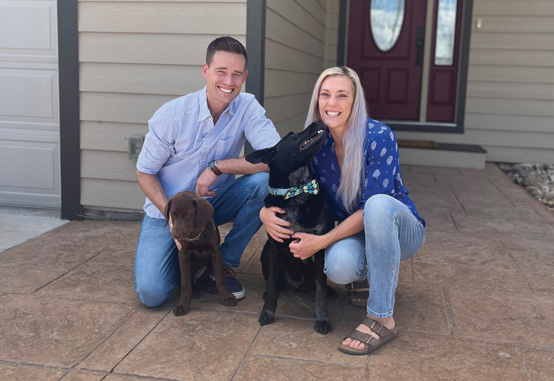 Two people kneeling in front of a house with a black dog