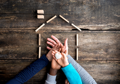 a pile of hands holding a white heart-shaped rock over a wood panel with a house made of blocks.