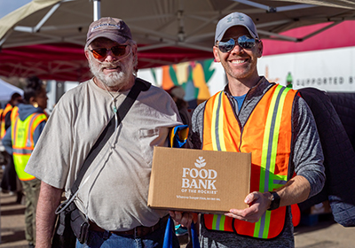 Two men - on a volunteer holding a box of food with Food Bank of the Rockies logo